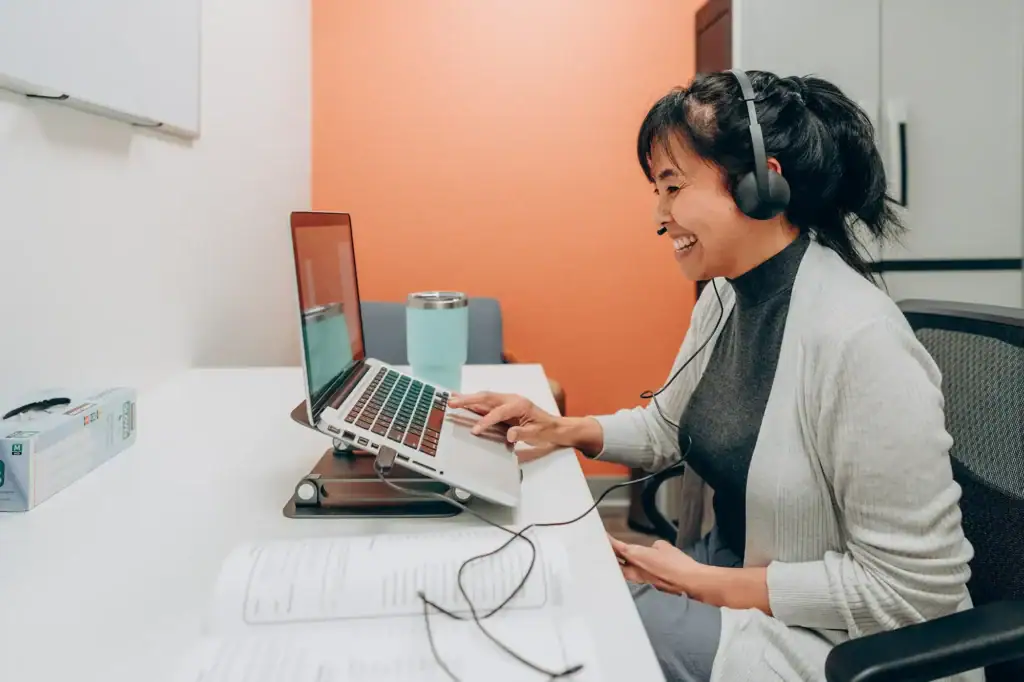 Telepractice speech therapy session with a smiling woman wearing headphones and working on a laptop for Speech Therapy Solutions Montana.